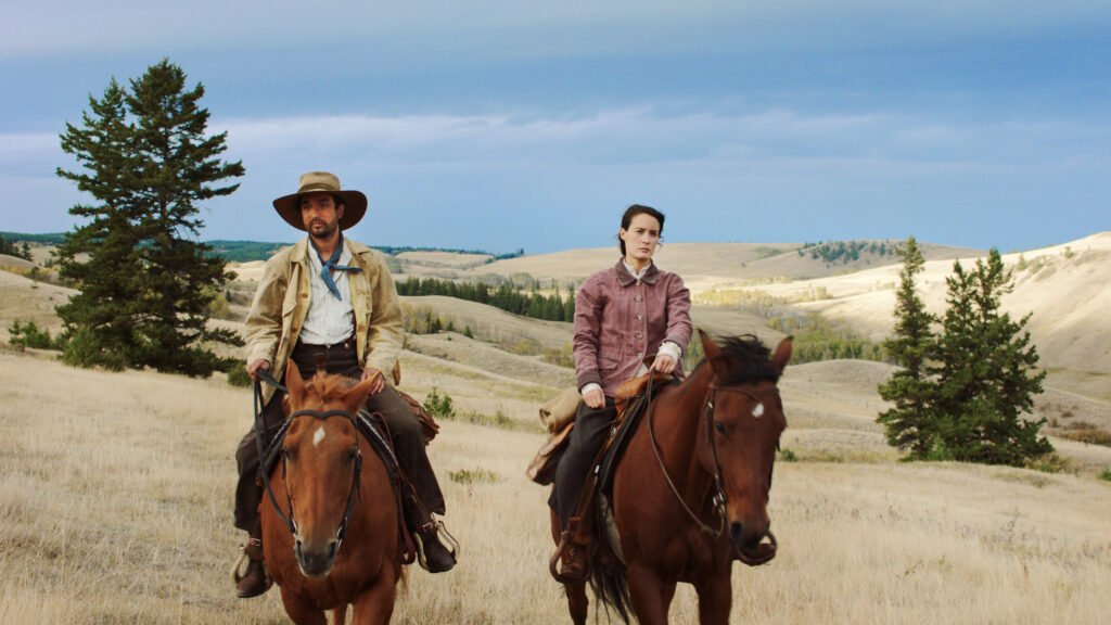 Two people on horseback ride through a vast, rolling landscape of hills and trees under a cloudy sky. Like an actress and director in an epic film, the rider on the left wears a large hat and beige jacket, while the one on the right dons a striking red jacket against the grassy terrain.