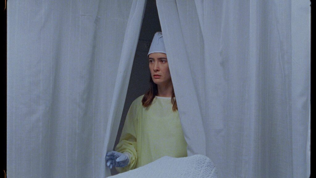 A woman in a yellow gown and cap, holding blue gloves, stands partially hidden by white curtains. She gazes intently forward, her expression serious. The scene suggests a medical or hospital setting, reminiscent of an actress preparing for her role under director Meredith Hama-Brown's guidance.