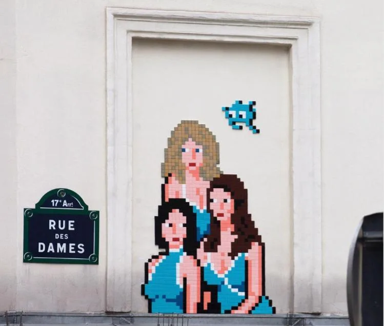 A pixel art mural by a renowned street artist adorns a building wall, depicting three women in turquoise dresses beside a small, blue pixel creature. A green street sign reading "Rue des Dames" reveals the 17th arrondissement location.
