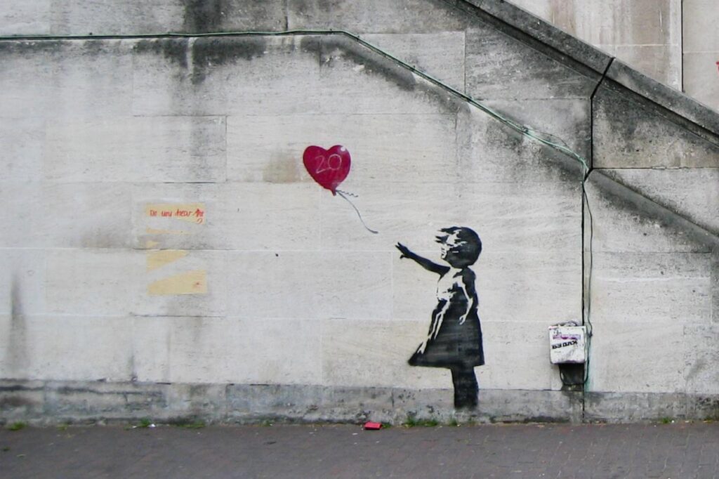 A graffiti artwork on a concrete wall, reminiscent of street artist Banksy's style, depicts a girl in a black dress reaching for a red heart-shaped balloon, marked with "20." The balloon floats away beside a large gray stone staircase, with some illegible text nearby.
