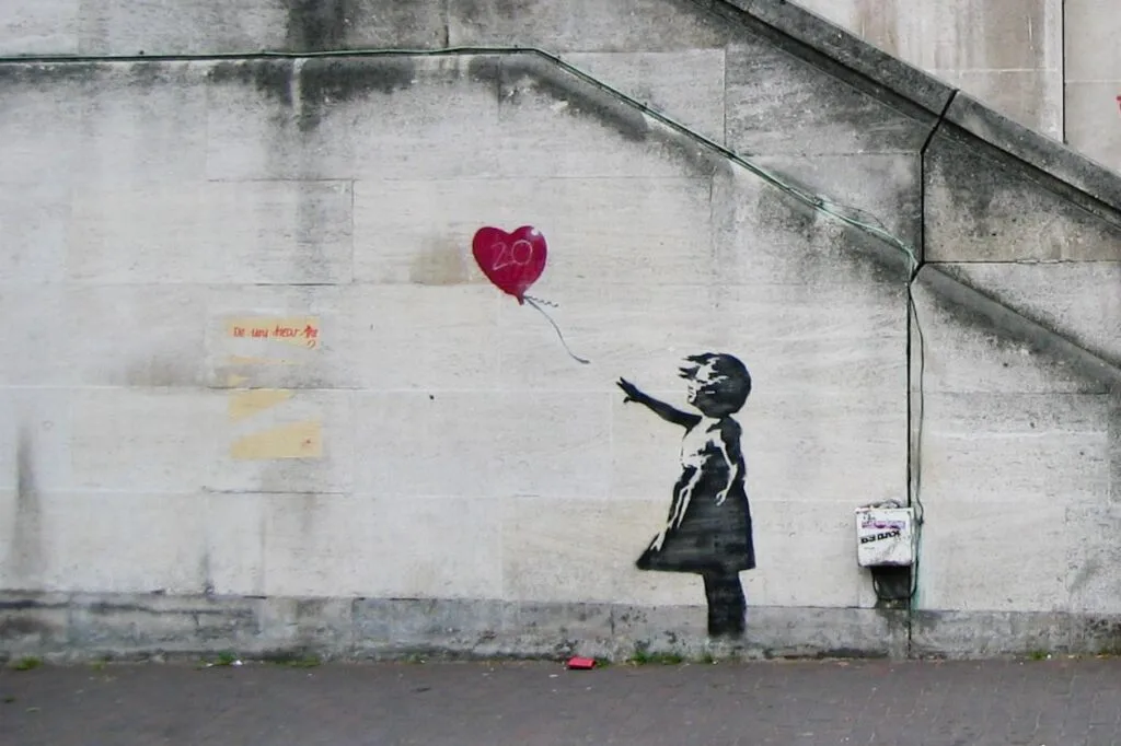 A graffiti artwork on a concrete wall, reminiscent of street artist Banksy's style, depicts a girl in a black dress reaching for a red heart-shaped balloon, marked with "20." The balloon floats away beside a large gray stone staircase, with some illegible text nearby.