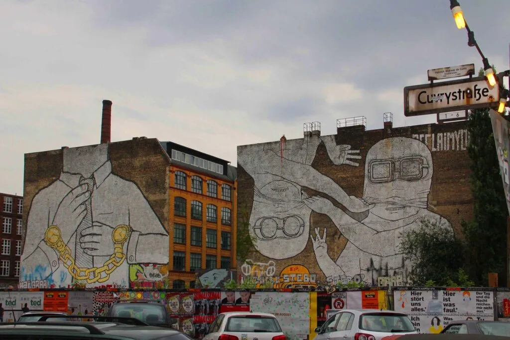A large brick building showcases two expansive murals reminiscent of street artists' work, featuring figures with exaggerated features. One wears a suit and gold chain, another gestures with an oversized hand, both in goggles. Below, vibrant graffiti and parked cars under a "Currystraße" sign capture the urban vibe.