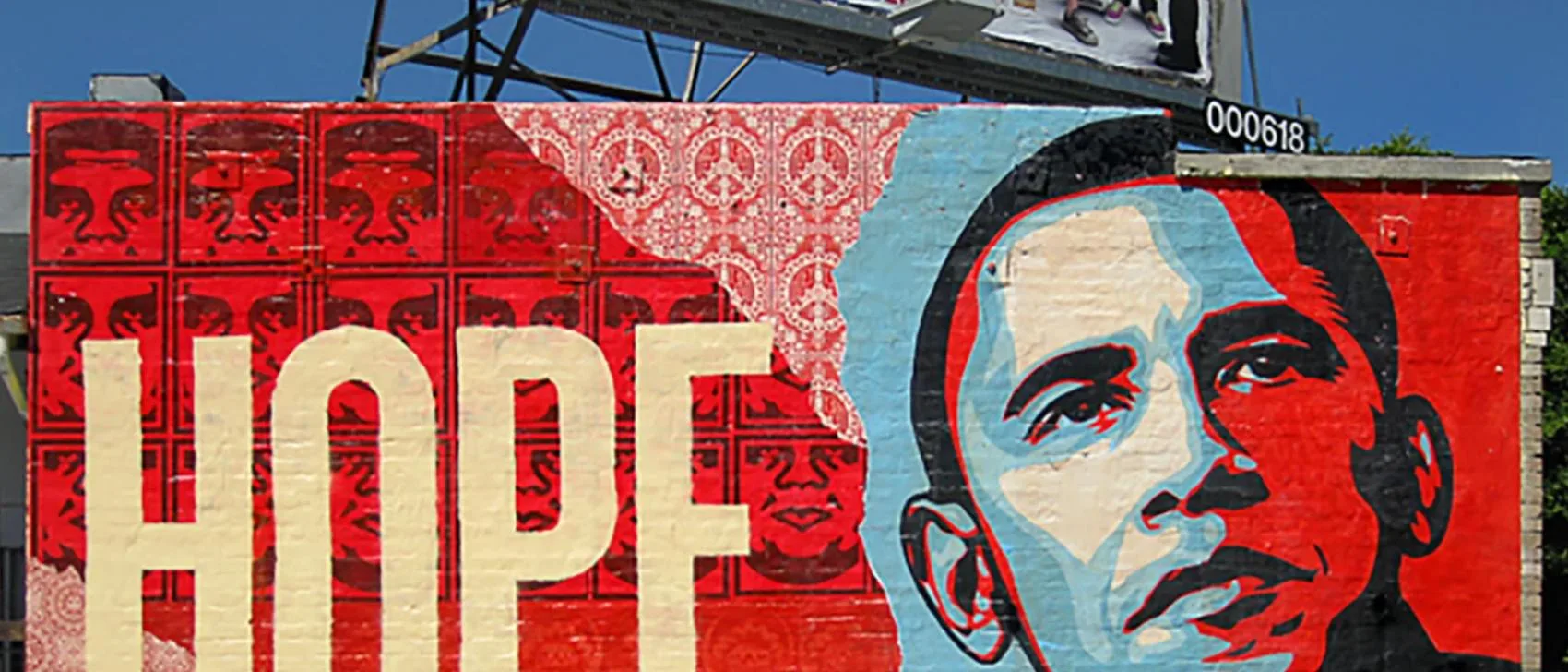 A colorful mural featuring stylized portraits on a red background and the word "HOPE" in bold cream letters echoes the spirit of street art. A large, blue-toned portrait reminiscent of Banksy's style is on the right. Above, a billboard with an unclear image stands against the clear blue sky.