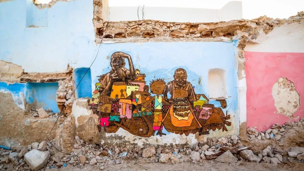 Street art on a crumbling wall depicts two figures adorned with colorful geometric patterns, set against a blue and pink painted background. Created by a renowned street artist, the mural is integrated into a dilapidated stone structure, surrounded by rubble and debris, suggesting an urban decay setting.