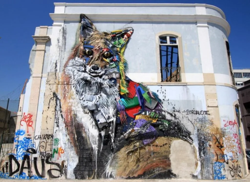 A vibrant street art mural on a weathered building depicts a colorful fox made of geometric shapes, channeling the creativity of renowned street artists. The fox is detailed with shades of red, blue, green, and yellow. Surrounding it are graffiti tags against the backdrop of broken windows.