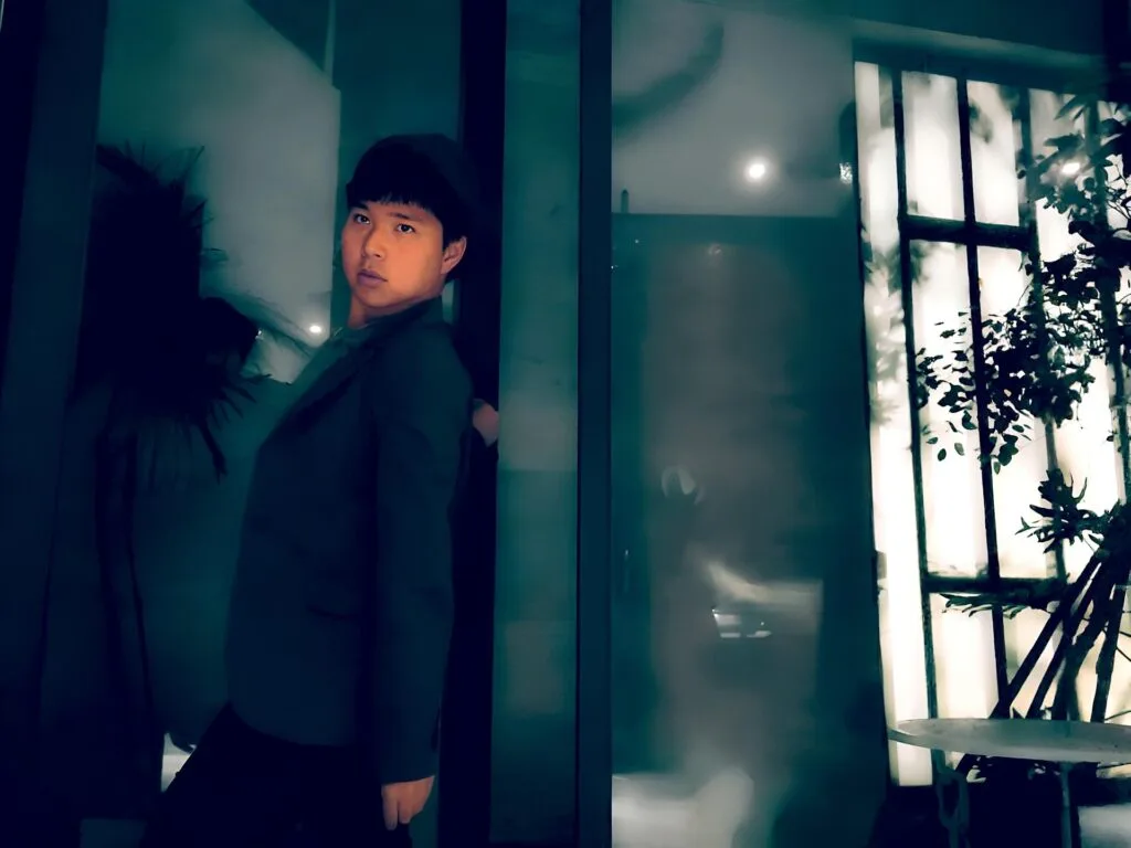 A person in a dark jacket leans slightly back against a teal glass wall, looking towards the camera. The background features dim lighting, blurred reflections, and silhouettes of plants, creating a moody ambiance.