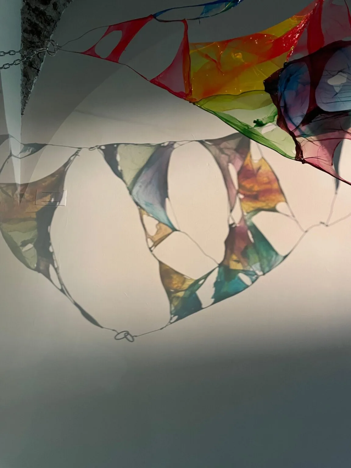 Colorful abstract installation by Giorgia Grassi with translucent panels in red, blue, yellow, and green shades, hanging on a wall. The pieces cast intricate shadows reminiscent of Simulacro d'infanzia, creating an interplay of light and color. Delicate shapes add depth and texture to the scene.