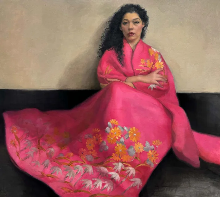 A person with long, curly hair wearing a pink robe adorned with vibrant floral designs sits on a dark bench against a muted background. The robe features yellow and white flowers, offering a contrast to the subdued setting. The person's expression is calm and contemplative.