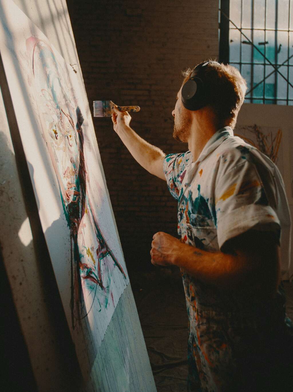 A man wearing headphones and a paint-splattered shirt paints an abstract portrait on a large canvas. Sunlight streams through a barred window, casting dramatic shadows, while the artist focuses intently on adding brushstrokes to the artwork.
