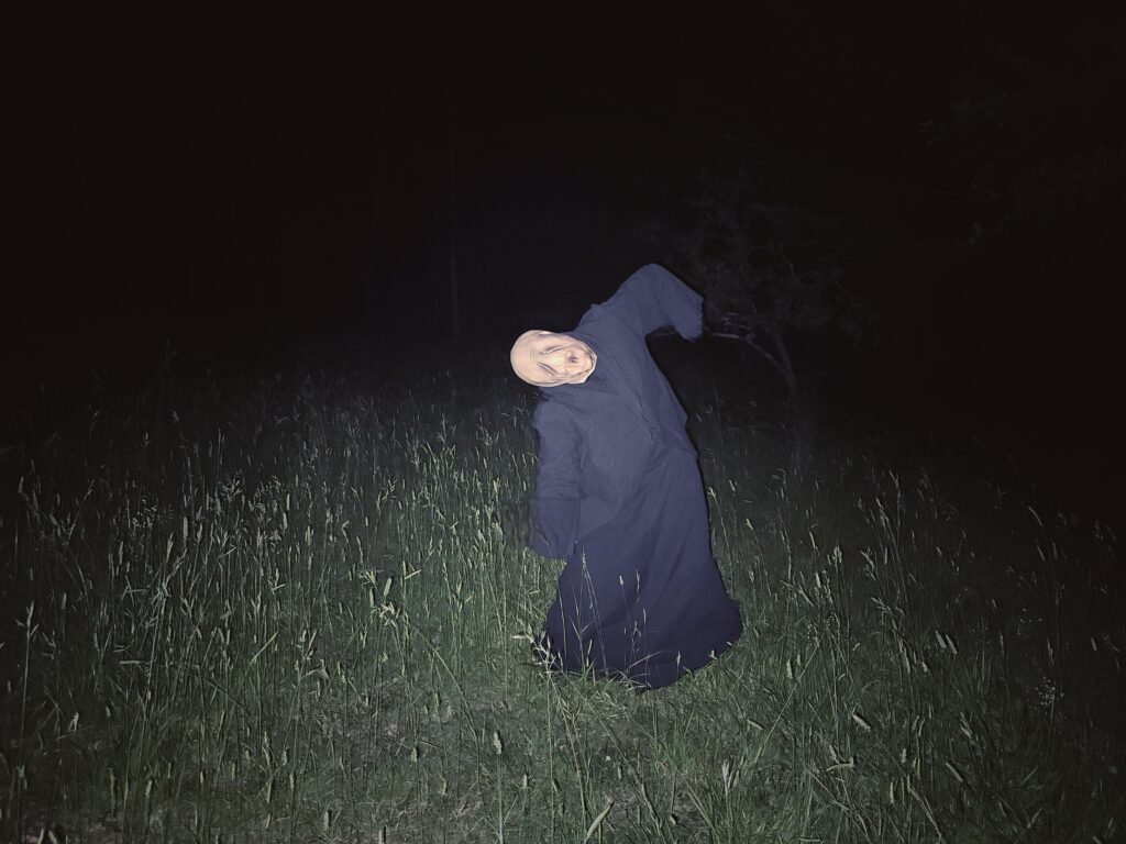 A person in a pale, eerie mask and long black robe stands in tall grass at night—part of a Gothic Multimedia Project. The figure’s head tilts unnaturally as a bright flash illuminates the scene, heightening the contrast with the darkness.