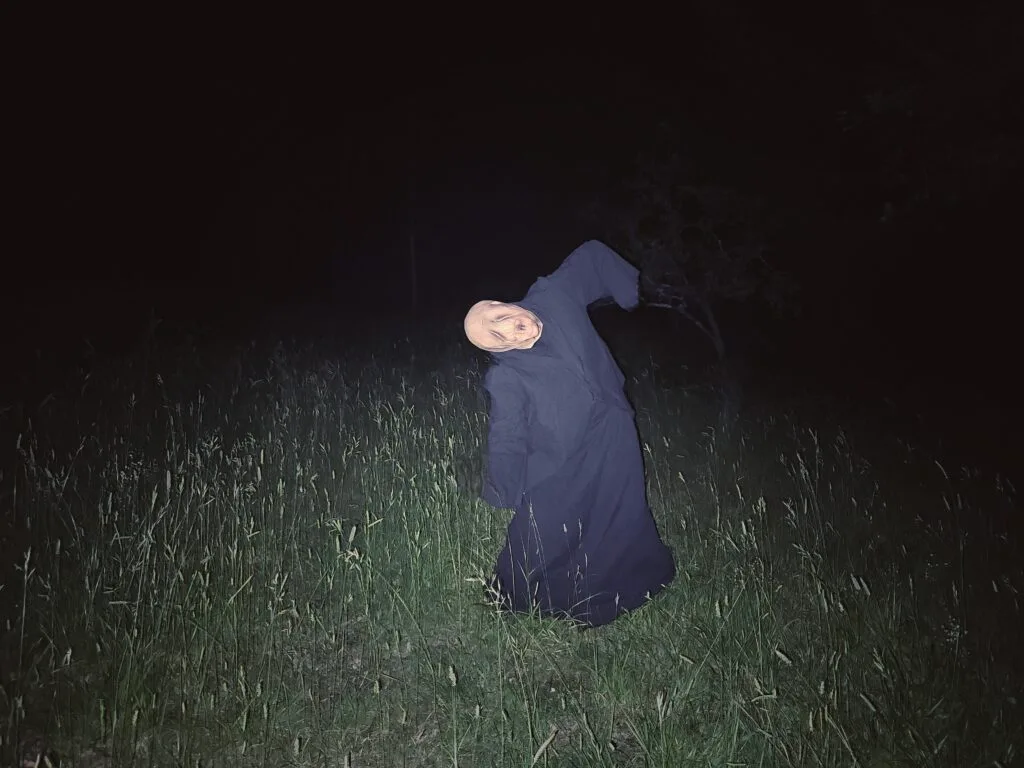 A person in a pale, eerie mask and long black robe stands in tall grass at night—part of a Gothic Multimedia Project. The figure’s head tilts unnaturally as a bright flash illuminates the scene, heightening the contrast with the darkness.