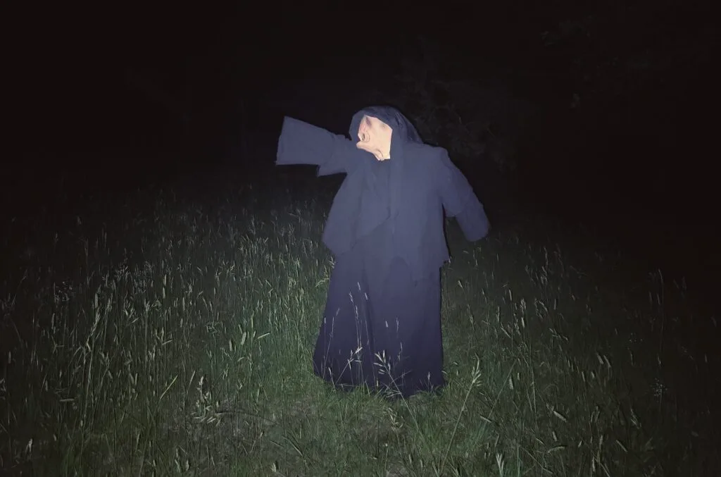 A figure in a long black robe and hood stands in tall grass at night. Their pale face is tilted upward, one arm raised as if reaching. The dim lighting heightens the eerie, mysterious atmosphere—perfect for a Gothic Multimedia Project.