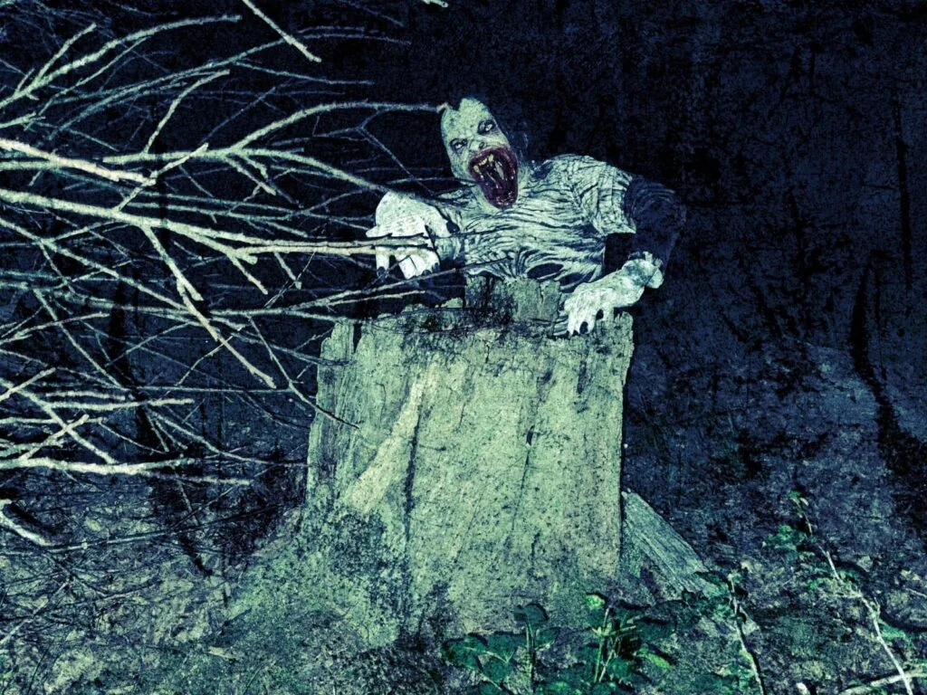 A creature with pale skin, dark sunken eyes, and a wide open mouth with sharp teeth emerges from behind a large tree stump at night—an eerie vision perfect for a Gothic Multimedia Project set in a shadowy forest framed by bare branches.