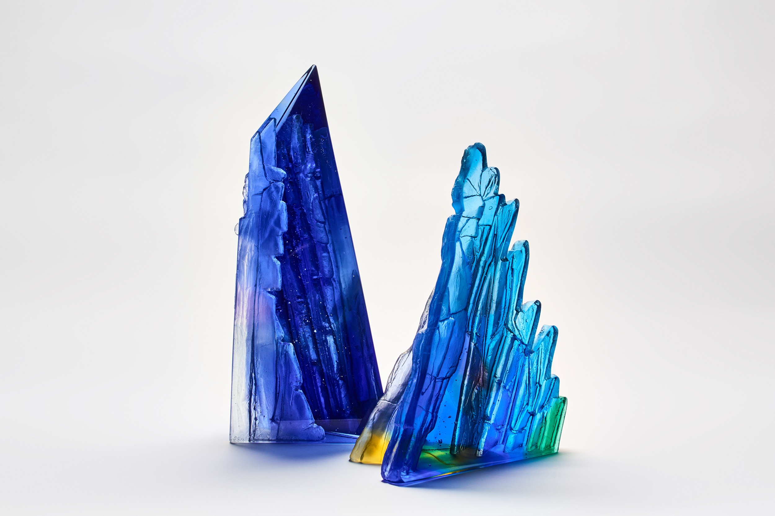 Two geometric glass sculptures resembling jagged crystals stand on a white background. The left structure is taller and deep blue, while the right is shorter, featuring blue, green, and yellow hues with sharp, angular edges. Both have a translucent, glossy finish.