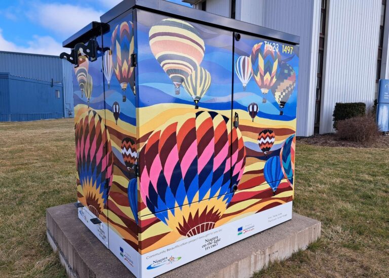 A utility box painted with colorful hot air balloons floating over rolling hills against a blue sky, placed on a concrete pad beside grass and a metal building. The artwork features vivid patterns and multiple balloons in various sizes.
